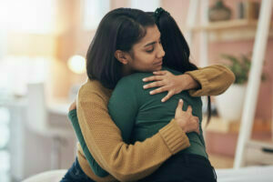 Picture of two young South Asian women hugging in a well lit room.  They both have long dark hair. One woman is wearing a green top and has her hair in a ponytail, and the other is wearing dark yellow. The woman in green has her face hidden form the camera.