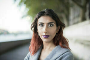 A transgender woman of West Asian/West Asian descent stands outside on a quiet city street. She is looking towards the camera with a neutral friendly expression. She is wearing purple lipstick and has shoulder length dark hair that has been dip dyed red. She is wearing a light grey jacket.