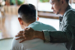 Rear view of a young Asian man and an older Asian man sitting together. The younger man is caring for the older one, putting hand on his shoulder, comforting and consoling him.